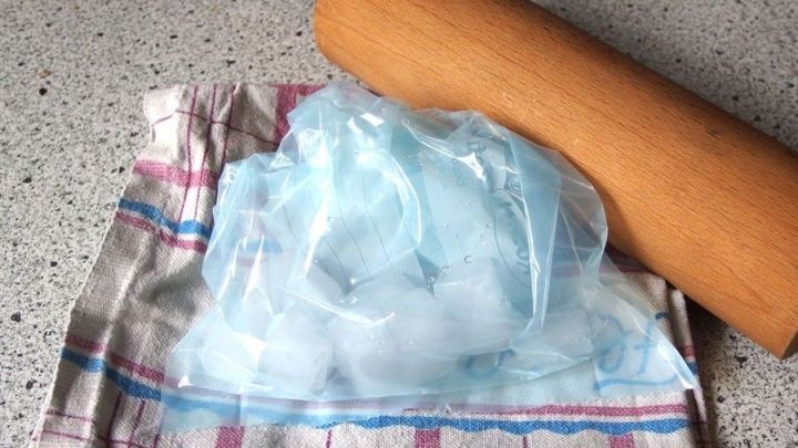 Eisw&uuml;rfel liegen in einem K&uuml;chentuch, w&auml;hrend ein Nudelholz zum Zerkleinern daneben bereitliegt.