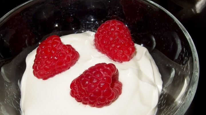 Ein Glas mit cremigem Quarkfondue, garniert mit frischen Himbeeren, steht auf einer dunklen Tischoberfl&auml;che.