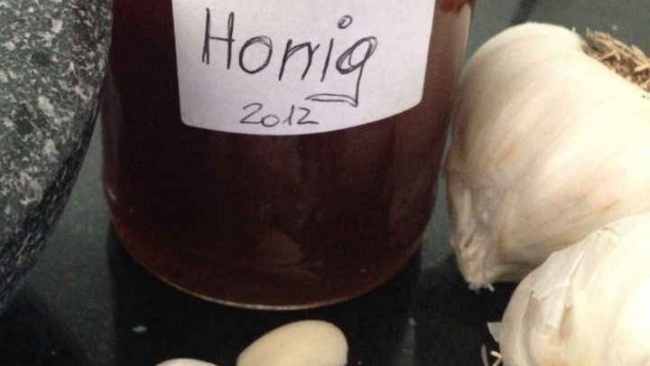 Ein Glas Honig steht auf einer Oberfl&auml;che, umgeben von frischem Knoblauch und einer Steinm&ouml;rser, was auf ein Heilmittel hinweist.