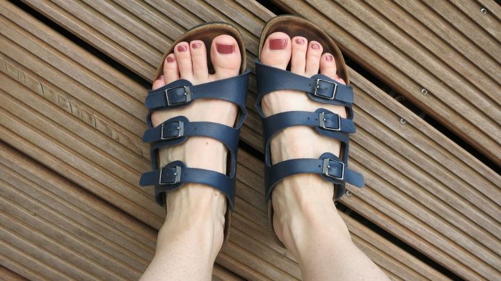 Raue F&uuml;&szlig;e liegen in blauen Sandalen auf einem Holzdeck; die F&uuml;&szlig;e sind gepflegt mit rotem Nagellack.