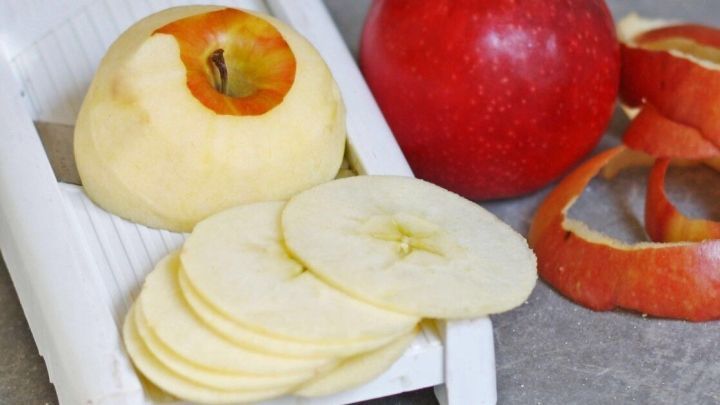 Scheiben von &Auml;pfeln liegen auf einer wei&szlig;en Hobel, w&auml;hrend ein roter Apfel und Apfelschalen daneben liegen.