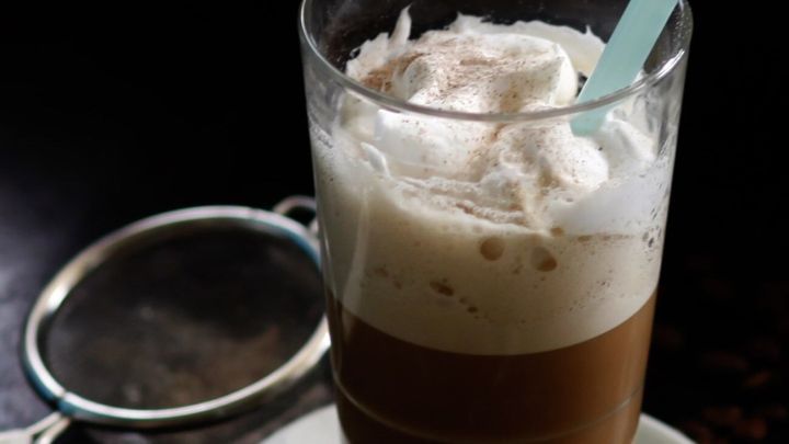 Ein Glas Kaffee-Frappe mit Sahnehaube und einem gr&uuml;nen Strohhalm steht auf einem Unterteller, umgeben von dunklem Hintergrund.