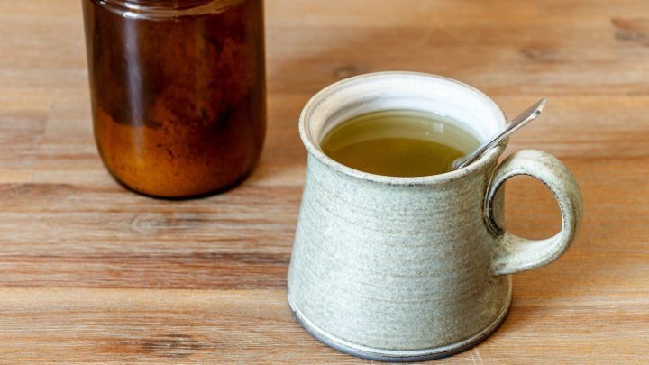Eine graue Tasse mit Br&uuml;he steht auf einem Holztisch, neben einem Glasbeh&auml;lter, der eine dunkle Fl&uuml;ssigkeit enth&auml;lt.
