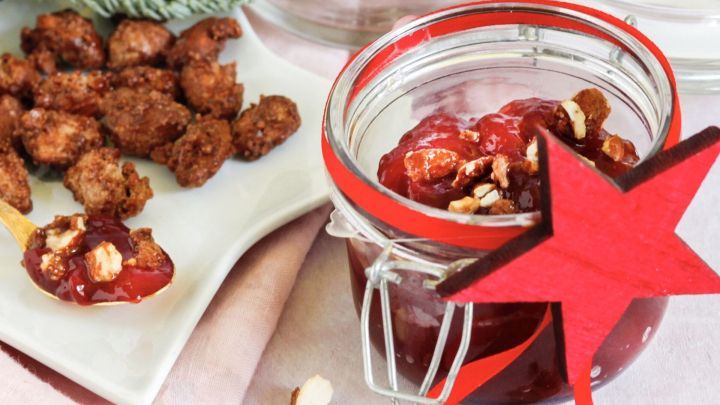 Ein Glas Himbeermarmelade mit Amaretto liegt auf einem Tisch, umgeben von gebrannten Mandeln und festlicher Dekoration.