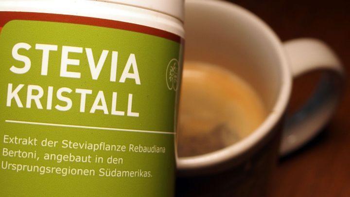 Eine Verpackung von Stevia Kristall steht neben einer Tasse Kaffee, die auf einem Holztisch steht, um Zuckerkonsum zu reduzieren.