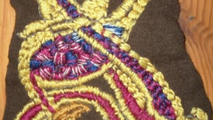 Ein buntes, abstraktes Stickmuster mit gelbem und rotem Garn auf braunem Stoff liegt auf einem Holztisch.