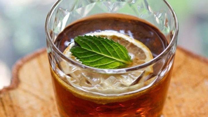 Ein Cocktailglas mit einer Zitronenscheibe und frischen Minzbl&auml;ttern steht auf einem Holztisch im Freien.