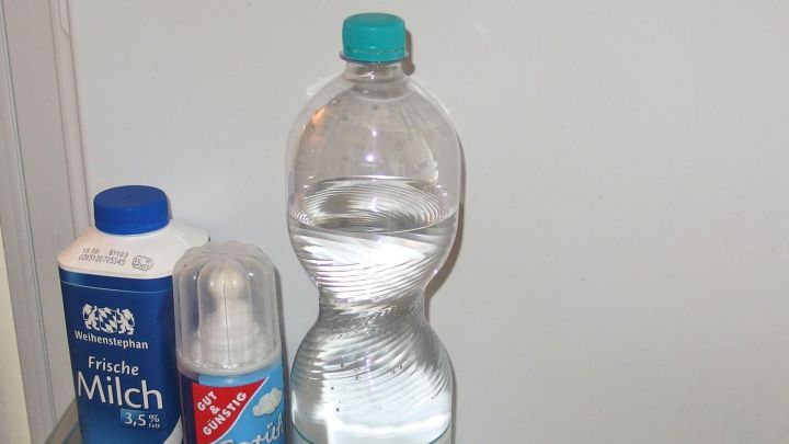 Eine Plastikflasche mit Wasser steht neben einer Milchpackung und einem Spray in einer hellen K&uuml;che, bereit zum Trinken.