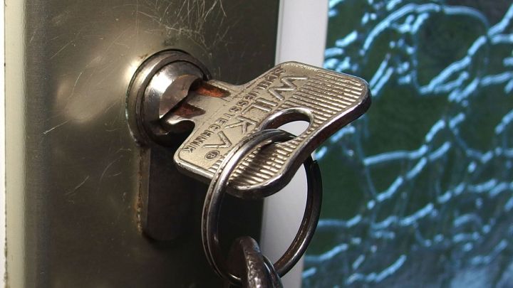 Ein Schl&uuml;ssel steckt im Schloss einer T&uuml;r, w&auml;hrend ein Schl&uuml;sselbund daneben h&auml;ngt, vor einer gl&auml;sernen Hintergrundfl&auml;che.