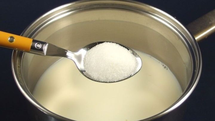 Eine Sch&ouml;pfkelle mit Zucker schwebt &uuml;ber einem Topf mit Milch, w&auml;hrend ein cremiger Pudding zubereitet wird.