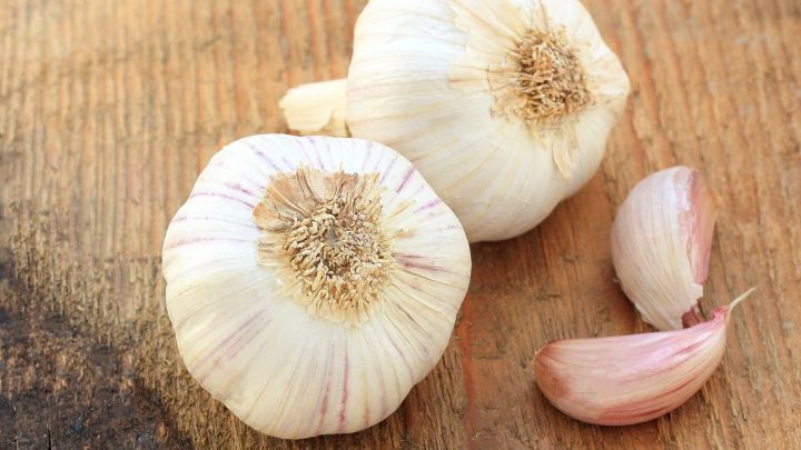 Zwei Ganze Knoblauchzehen liegen auf einem Holzbrett, w&auml;hrend eine gesch&auml;lte Knoblauchzehe daneben platziert ist.