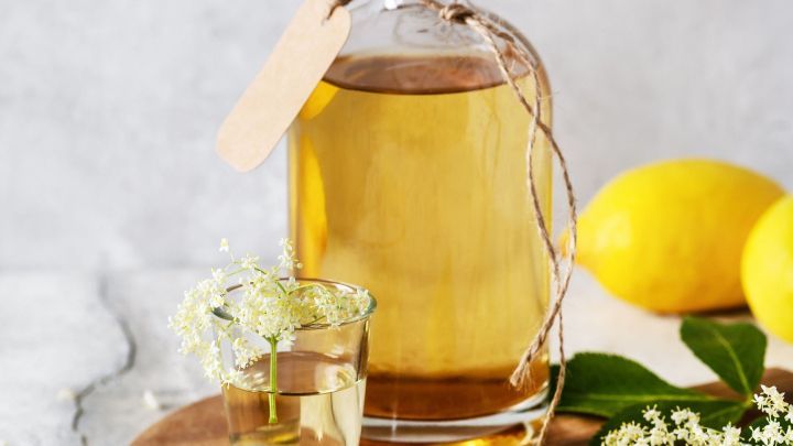 Eine Flasche Holunderbl&uuml;tenschnaps steht auf einem Holzbrett, umgeben von Zitronen und Holunderbl&uuml;ten, w&auml;hrend ein Glas gef&uuml;llt ist.