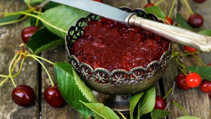 Ein gl&auml;nzendes Marmeladenglas steht auf einer rustikalen Holzfl&auml;che, umgeben von frischen Kirschen und Bl&auml;ttern.
