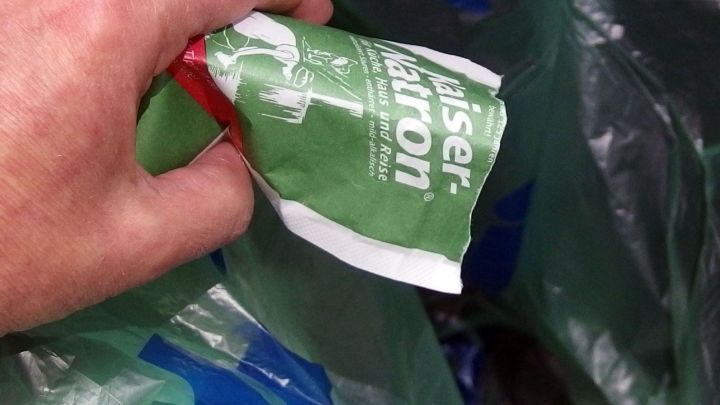 Eine Hand h&auml;lt ein gr&uuml;nes P&auml;ckchen eines Geruchsneutralisierers, w&auml;hrend sie Papier- und Plastikt&uuml;ten in einem M&uuml;lleimer umschichtet.
