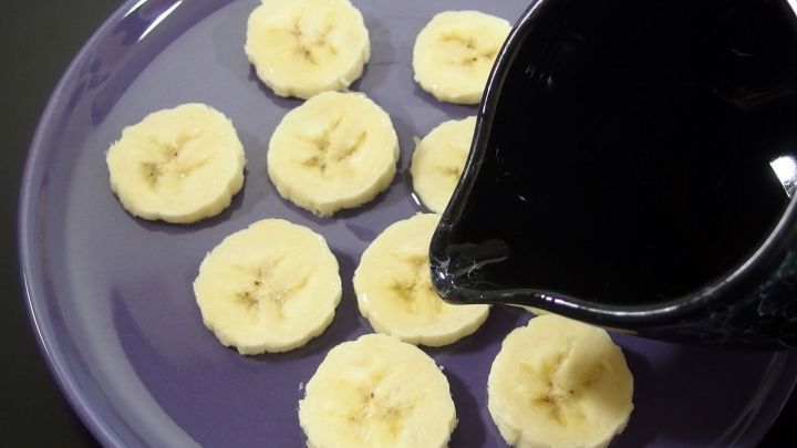 Gefrorene Bananenscheiben liegen auf einem lila Teller, w&auml;hrend eine schwarze Kanne Lik&ouml;r dar&uuml;ber gegossen wird.