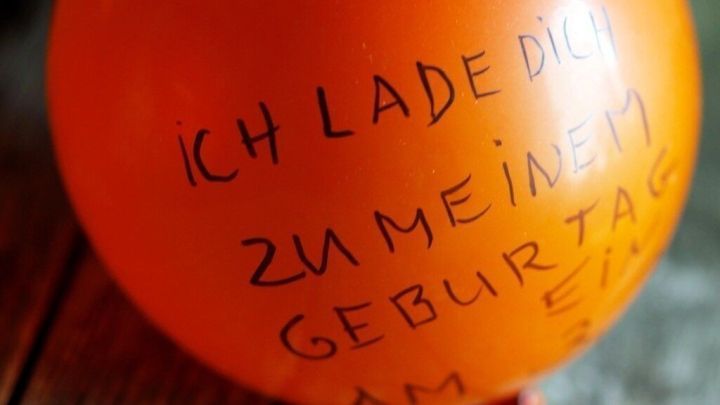 Ein leuchtend orangefarbener Ballon steht auf einem rustikalen Holztisch, darauf steht: &bdquo;ICH LADE DICH ZU MEINEM GEBURTSTAG AM&hellip;&ldquo;.