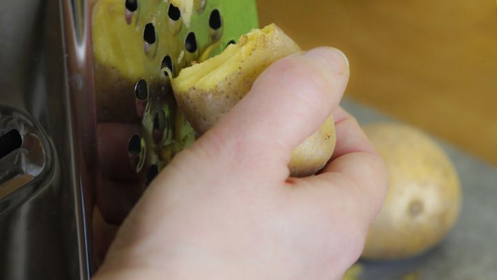 Eine Hand reibt eine Kartoffel an einer Reibe, w&auml;hrend weitere Kartoffeln auf der Fl&auml;che liegen, um Hausmittel gegen geschwollene Augen zuzubereiten.