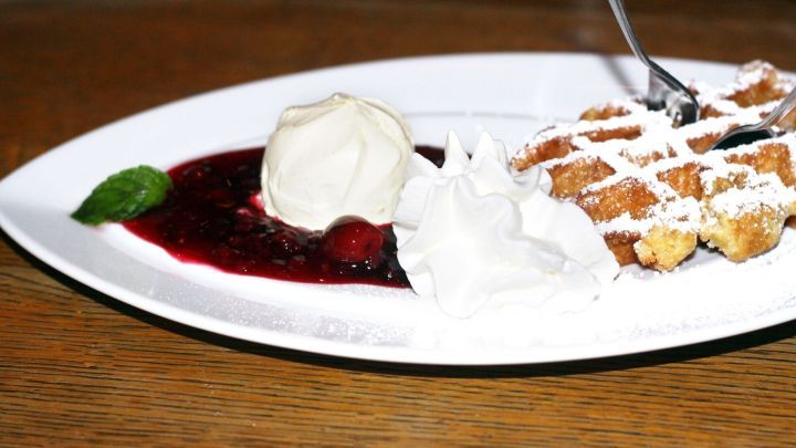 Fluffige Waffeln liegen auf einem Teller, umgeben von Sahne, roter Fruchtso&szlig;e und einem Minzblatt in einer rustikalen Umgebung.