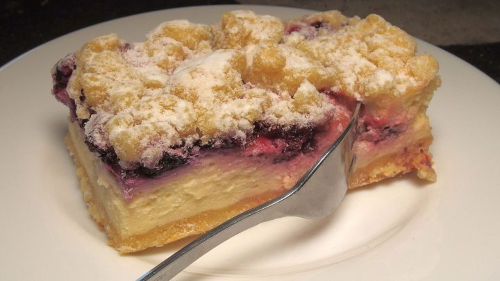 Ein St&uuml;ck K&auml;sekuchenhilfe liegt auf einem wei&szlig;en Teller, w&auml;hrend ein L&ouml;ffel daneben zum Servieren bereitsteht.