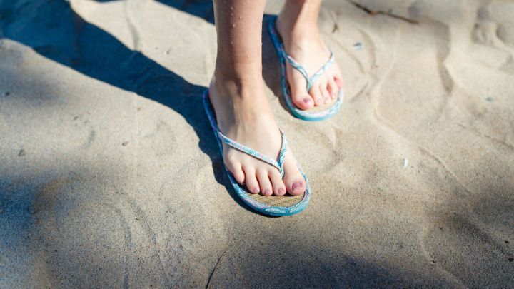 Flip-Flops liegen im Sand, w&auml;hrend eine Person mit nackten F&uuml;&szlig;en darin steht und sich m&ouml;glicherweise leicht unwohl f&uuml;hlt.