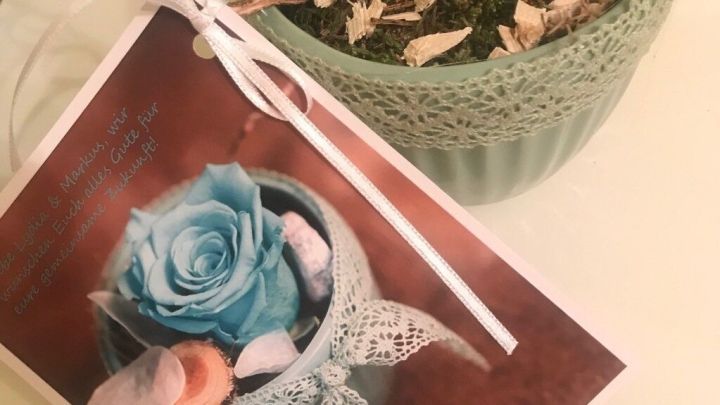 Eine dekorative, t&uuml;rkisfarbene Rose in einem Topf pr&auml;sentiert sich neben einem Geschenkanh&auml;nger, der das Arrangement beschreibt.