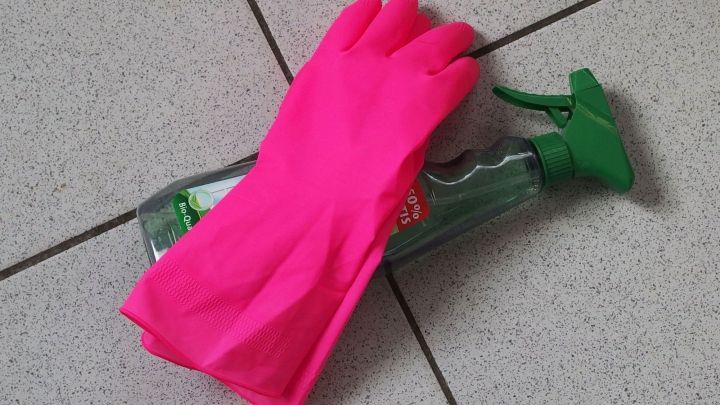 Eine pinke Gummihandschuh liegt auf einer Flasche Putzmittel, umgeben von grauen Fliesen in einem Reinigungsumfeld.