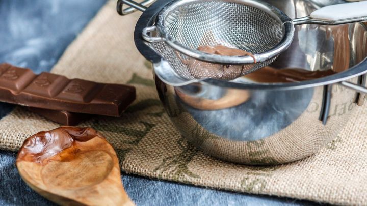 Ein metallischer Sieb ist &uuml;ber einer Sch&uuml;ssel platziert, w&auml;hrend geschmolzene Schokolade und ein Holzl&ouml;ffel daneben liegen.
