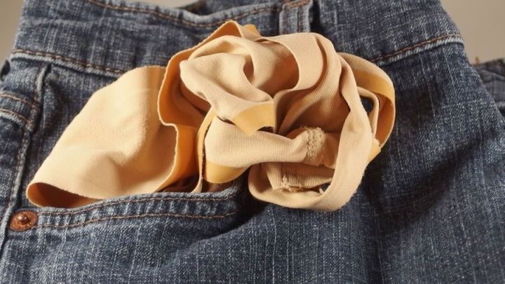Ein Kleidungsst&uuml;ck mit einem beige Stofffetzen steckt in der Tasche einer Jeans, die auf einem neutralen Hintergrund liegt.