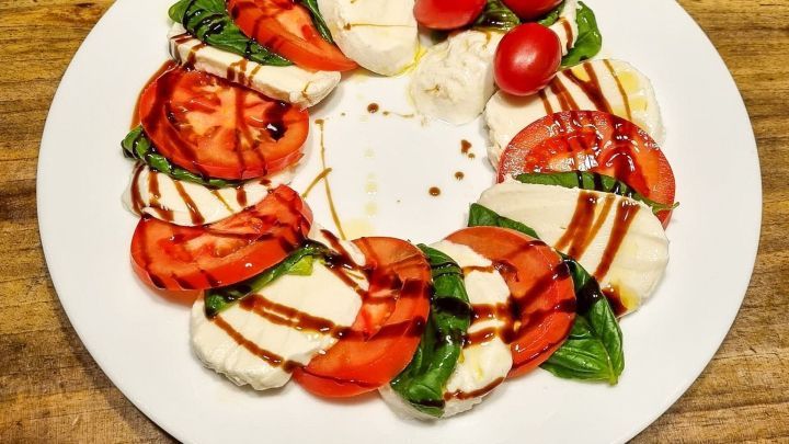 Auf einem wei&szlig;en Teller liegt eine kunstvolle Anordnung aus Tomaten, Mozzarella und frischem Basilikum, garniert mit Balsamico.
