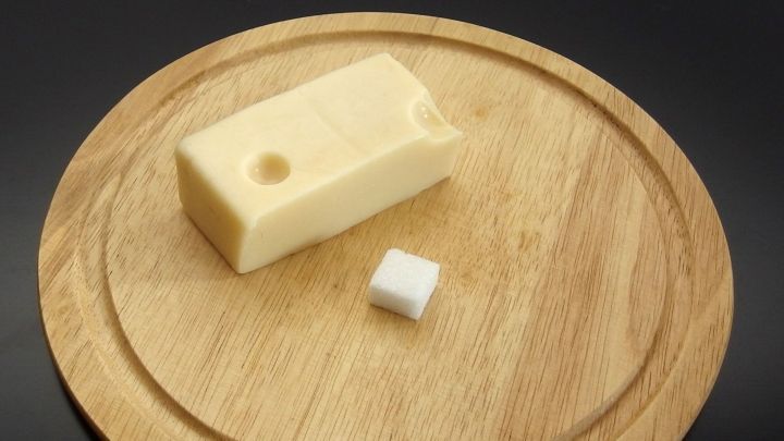 Ein St&uuml;ck K&auml;se liegt auf einem Holzbrett neben einem W&uuml;rfel Zucker, der zur Frischhaltung verwendet wird.