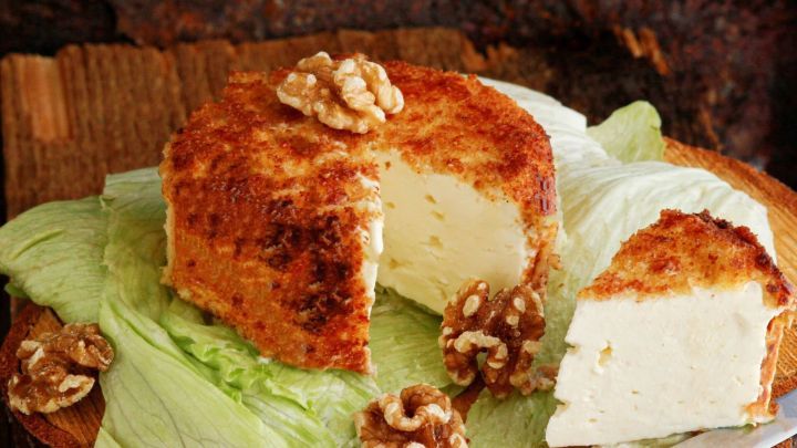 Paniert Feta-K&auml;se liegt auf gr&uuml;nem Blatt-Salat, umgeben von Waln&uuml;ssen, auf einem Holzbrett mit einem Messer.