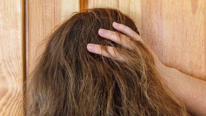 Ein Hand greift durch fettige Haare, w&auml;hrend der Hintergrund aus einer Holzwand besteht, was eine ungepflegte Umgebung suggeriert.