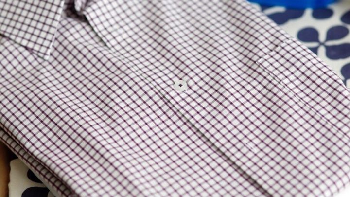 Ein kariertes Hemd liegt ordentlich gefaltet auf einem B&uuml;gelbrett, neben einem blauen B&uuml;geleisen auf einem gemusterten Stoff.