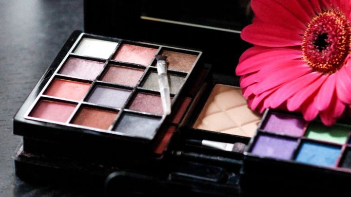 Ein Kosmetik-Set mit Lidschattenfarben liegt offen auf einem dunklen Tisch, erg&auml;nzt von einer pinken Blume.