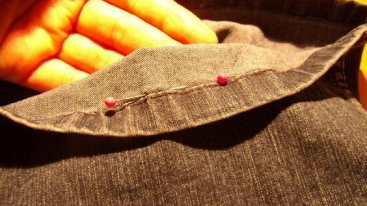 Eine Hand h&auml;lt Jeansstoff, der mit Stecknadeln gesichert ist, um den Original-Saum korrekt zu k&uuml;rzen.