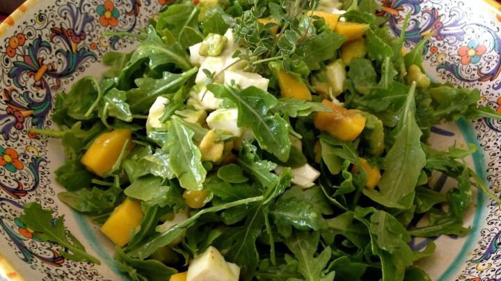 Ein frischer Avocado-Mozzarella-Salat liegt ansprechend in einer bunten Sch&uuml;ssel, umgeben von Mango- und Rucola-St&uuml;cken.