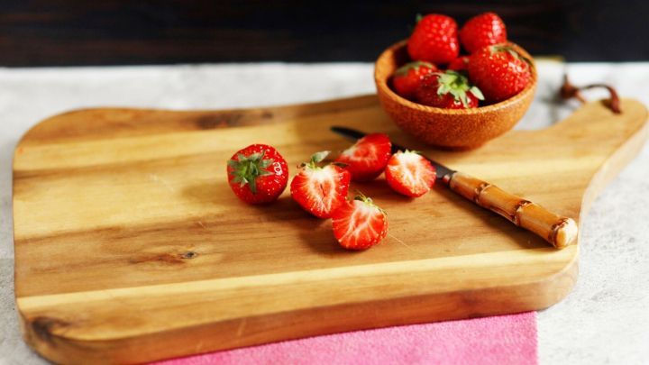 Ein Holzbrett liegt auf einer Arbeitsfl&auml;che, w&auml;hrend frische Erdbeeren und ein Messer darauf liegen. Ein Sch&uuml;sselchen mit Erdbeeren steht daneben.