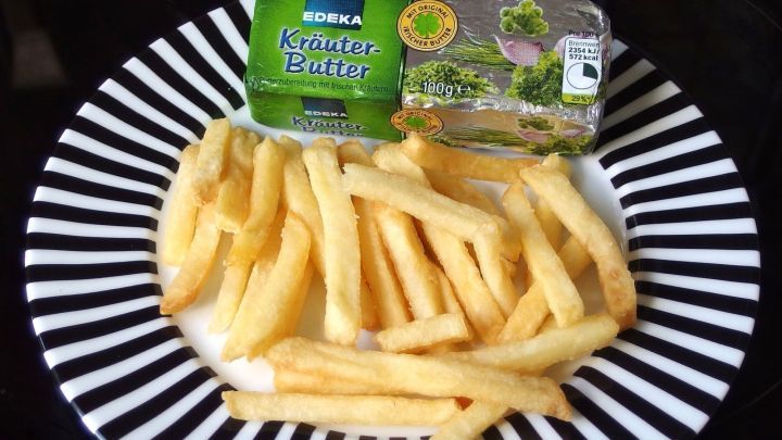 Pommes liegen auf einem schwarz-wei&szlig;en Teller neben einem kleinen Block Kr&auml;uterbutter. Sie wirken knusprig und einladend.