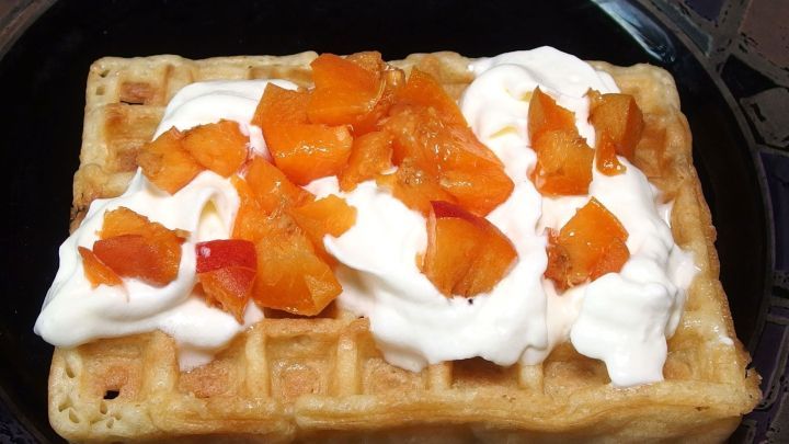 Eine Waffel liegt auf einem schwarzen Teller, garniert mit Sahne und bunten Fruchtst&uuml;cken, in einer einladenden K&uuml;chenszene.