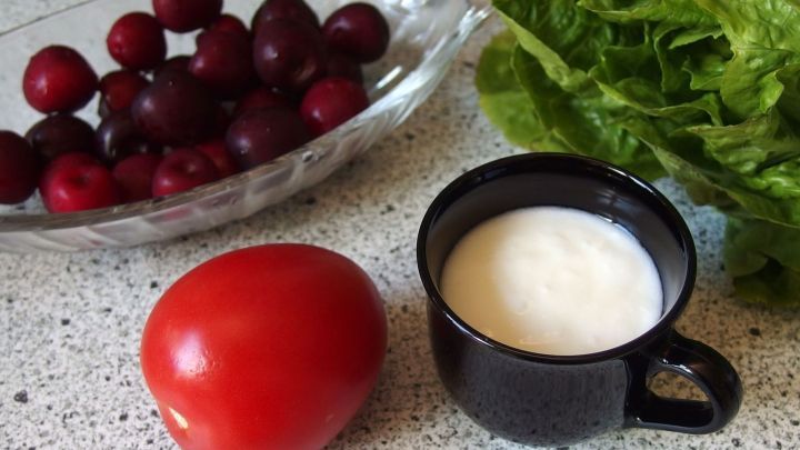 Eine rote Tomate liegt neben einer schwarzen Tasse mit einer Creme, umgeben von frischem Salat und Trauben auf einer Arbeitsfl&auml;che.