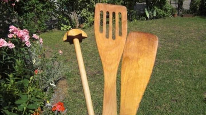 Holzl&ouml;ffel und Pfannenwender trocknen in der Sonne, gehalten von einer Hand, im Garten mit gr&uuml;ner Wiese und bl&uuml;henden Pflanzen.