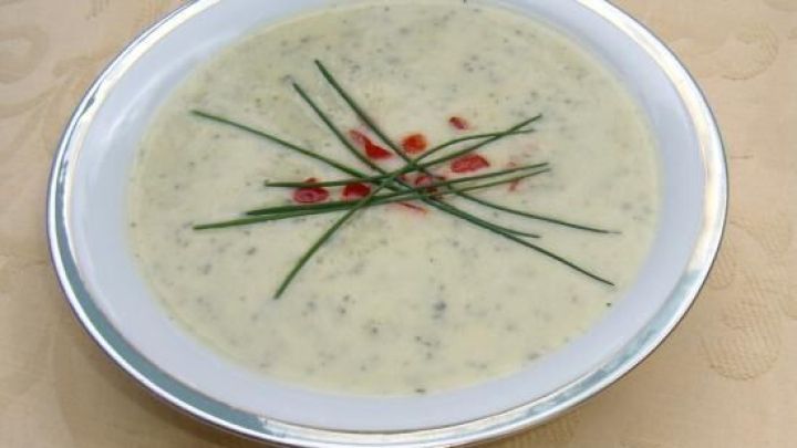 Eine cremige Zucchini-Cremesuppe in einer wei&szlig;en Sch&uuml;ssel, verziert mit Kr&auml;utern und roten Punkten, auf einem eleganten Tisch.