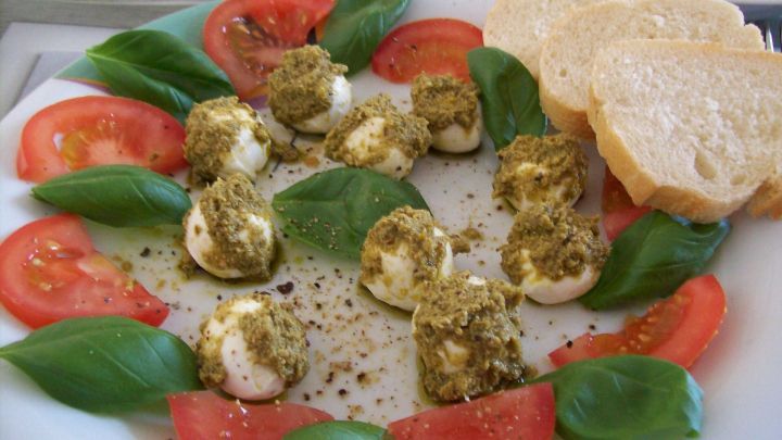 Mozzarellaw&uuml;rfel mit Pesto auf Teller, umrahmt von frischen Basilikumbl&auml;ttern und Tomatenscheiben, daneben Brotscheiben.