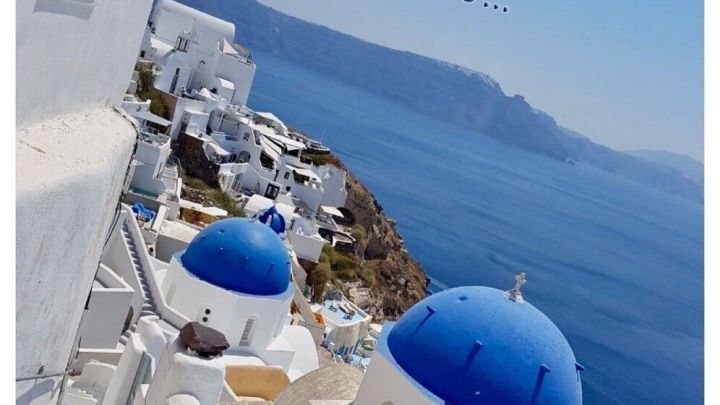 Eine idyllische Aussicht zeigt wei&szlig;e H&auml;user mit blauen D&auml;chern, w&auml;hrend der Text "Liebe Oma, hier ist's sch&ouml;n..." vermerkt ist.