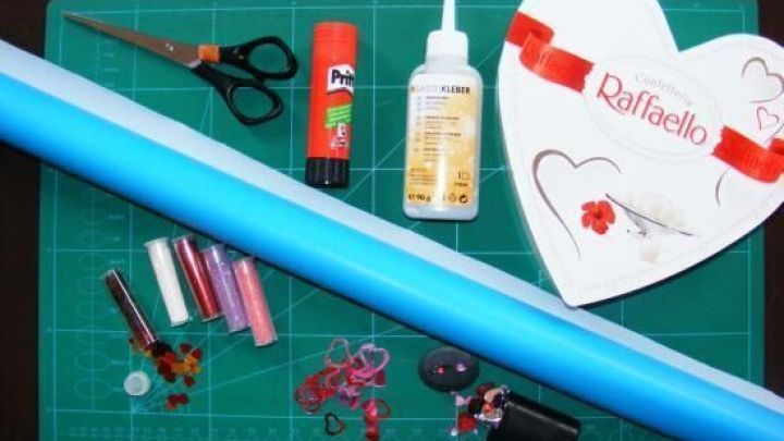 Eine herzf&ouml;rmige Raffaello-Schachtel liegt auf einem gr&uuml;nen Bastelbrett, umgeben von Bastelmaterialien wie Scheren, Klebstoff und Dekos.