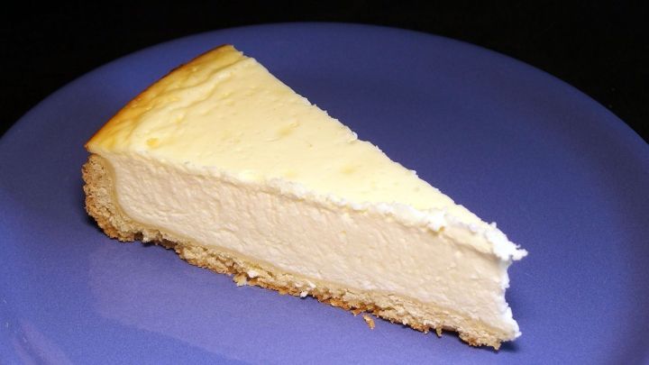 Ein St&uuml;ck K&auml;sekuchen liegt auf einem blauen Teller, bereit zum Genie&szlig;en in einer dunklen Umgebung.