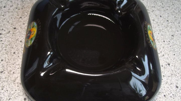 Ein schwarzer Aschenbecher liegt auf einer hellen, gemusterten Oberfl&auml;che und scheint unbenutzt zu sein.