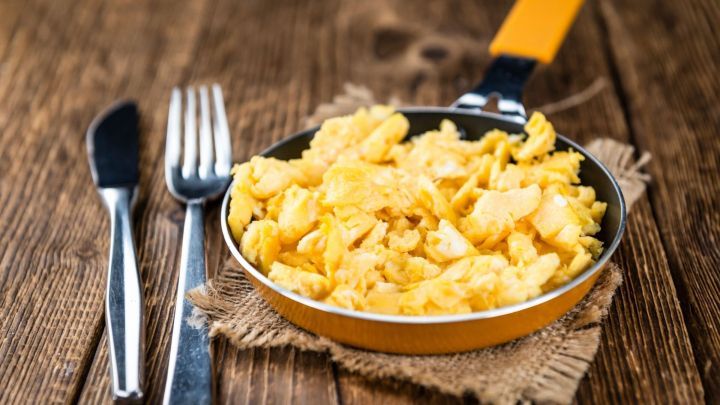 Eine kleine Pfanne mit bunten R&uuml;hreiern steht auf einem rustikalen Holztisch, umgeben von Besteck.