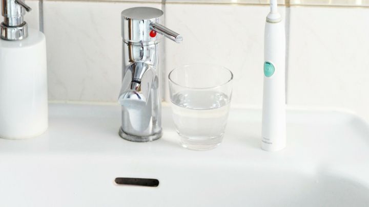 Ein elektrischer Zahnb&uuml;rste steht neben einem Glas Wasser auf einem Waschbecken, umgeben von Pflanzen in goldenen T&ouml;pfen.