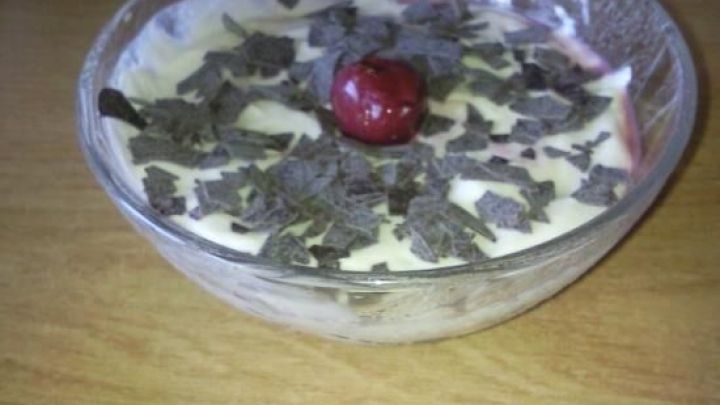 Eine Glasschale zeigt einen Schwarzwaldbecher, bedeckt mit Schokoraspeln und einer Kirsche, auf einem Holztisch stehend.